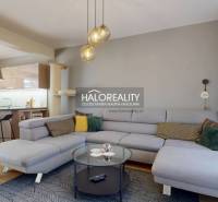 Prostorný obývací pokoj v rodinném domě s pohovkou, kuchyňským koutem a podlahou s dřevěným dekorem.