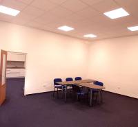 Kancelář s modrými židlemi a stoly, bílá stěna, otevřené dveře k malé kuchyňce.