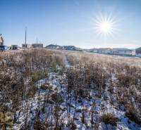 Sněhem pokrytý pozemek v Mlynici s obydlími v pozadí, ideální pro bydlení.