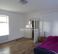 Pokoj s postelí, oknem, podlahou s dřevěným dekorem, nočním stolkem a otevřenými dveřmi.