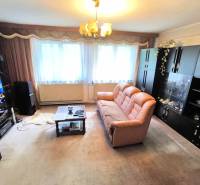 Obývací pokoj v rodinném domě s hnědou pohovkou, skříňkami a televizí na stěně.