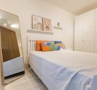 Ložnice v jednopokojovém bytě s postelí, barevnými polštáři, zrcadlem a velkou skříní.