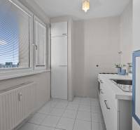 Kuchyně v 1-pokojovém bytě s bílým nábytkem a kachličkami, s výhledem přes žaluzie.