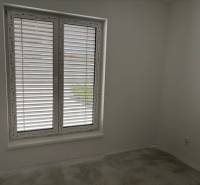 Pokoj v rodinném domě s velkým oknem a žaluziemi, bílé stěny, šedá podlaha.