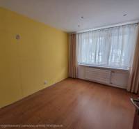 Pokoj se žlutou stěnou, oknem s jemnými záclonami a podlahou s dřevěným dekorem.