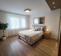 Ložnice s postelí, zrcadlem a nočními stolky ve 4pokojovém bytě s podlahou s dřevěným dekorem.