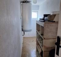 Úzký interiér rodinného domu s bojlerem a poličkami, světlé stěny, podlaha s dřevěným dekorem.