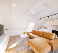 Obývací pokoj s kuchyní ve 2pokojovém bytě: velké okno, hnědá sedačka, podlaha s dřevěným dekorem.