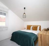 Ložnice v rekreačním apartmánu s manželskou postelí, oknem a komodou pod šikmou střechou.