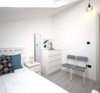 Rekreační apartmán se světlou ložnicí, postelí, komodou a polštáři, podlaha s dřevěným dekorem.