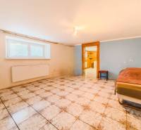 Pokoj s dlažbou v rodinném domě, postel u stěny, okno s radiátorem.