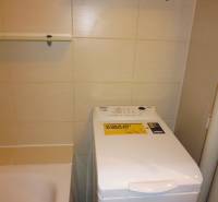 Koupelna v 2-pokojovém bytě s pračkou vedle vany a policemi.