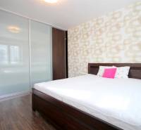 Ložnice v dvoupokojovém bytě s postelí, vestavěnou skříní a podlahou s dřevěným dekorem.