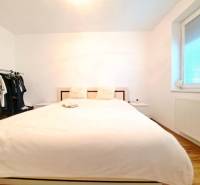 Ložnice ve dvoupokojovém bytě s postelí, otevřenou šatní skříní a podlahou s dřevěným dekorem.