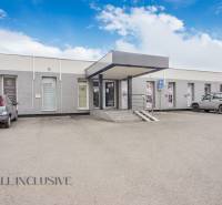 Jednopodlažní budova s obchodními prostory na ulici Vajanského, parkovištěm a auty před vstupem.