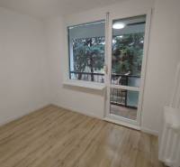 Pokoj se světlými stěnami, podlahou s dřevěným dekorem a výhledem na zeleň.