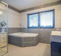 Koupelna v rodinném domě s vanou, sprchou a umyvadlem, světlé obklady, okno s roletami.