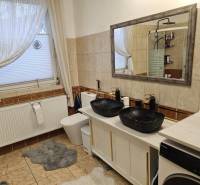 Koupelna v rodinném domě s dvojitým umyvadlem, vanou a pračkou na dlažbě.