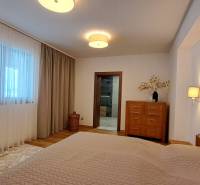 Ložnice v dvoupokojovém bytě s dřevěným dekorem, závěsy a světlým nábytkem.