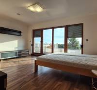 Ložnice v rodinném domě s podlahou s dřevěným dekorem, postelí a velkými balkonovými okny.