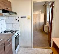 Kuchyně s bílými obkladačkami, sporákem, skříňkami a pohled do předsíně v 1-pokojovém bytě.