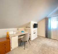 Pokoj v rodinném domě s pracovním stolem, televizí a světlým kobercem.