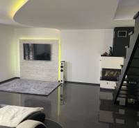 Obývací pokoj v rodinném domě se schodištěm, krbem a televizí na stěně.