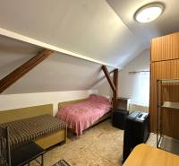 Podkrovní pokoj v rodinném domě s jednoduchým nábytkem a střešními trámy.