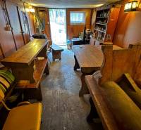 Interiér chaty s dřevěnými stoly a lavicemi, výhled na zahradu.