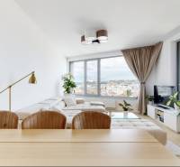 Obývací pokoj třípokojového bytu se světlou sedací soupravou, velkými okny a výhledem na město.