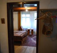 Interiér rodinného domu s postelí a dřevěnou podlahou, přes dveře je vidět pokoj.