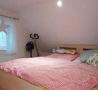 Ložnice v podkroví rodinného domu s postelí, televizorem a podlahou s dřevěným dekorem.