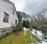 Zahrada u chaty v Modre s trávníkem, sněhem a dřevinami podél domu.