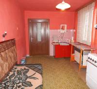 Kuchyně v rodinném domě s červenými stěnami, starším nábytkem a zeleným kobercem.