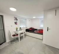 Interiér chaty s jídelním stolem, židlemi a postelí na světlé podlaze.