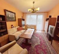 Obývací pokoj v 2-pokojovém bytě se starším nábytkem, kobercem a dekorativními závěsy.