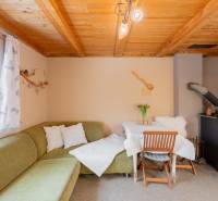 Interiér chaty s kachlovými kamny, zelenou sedačkou a stolem. Dřevěný strop a dekorace.