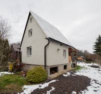 Chata v Handlové s trávníkem a zasněženou zahradou, obklopená stromy a keři.