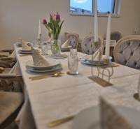 Stůl s bílým ubrusem, talíře, sklenice, květiny a potahy židlí v gastro prostorech.