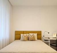 Ložnice v jednopokojovém bytě se světlou postelí, záclonami a bílým stolem s lampou.