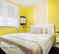 Ložnice v rodinném domě se žlutými stěnami, bílou postelí a květinovou dekorací.