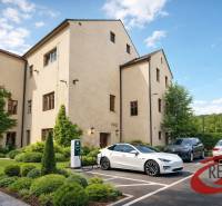 Budova ve Velkých Uhercích se zelení a nabíječkou pro elektrická auta na parkovišti.