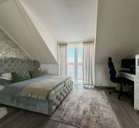 Ložnice v dvoupokojovém bytě s manželskou postelí, psacím stolem a podlahou s dřevěným dekorem.