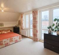 Ložnice v rodinném domě s postelí, skříňkou, velkými okny a rostlinami na parapetu.