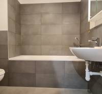 Koupelna s vanou, umyvadlem a pračkou v 2-pokojovém bytě. Šedé obklady dominují prostoru.