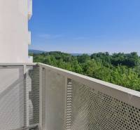 Balkón s výhledem na les a kopce, budova v pozadí, jasná obloha.