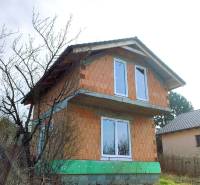 Dvoupatrová chata v rozestavěném stavu, obklopená stromy a trávou na venkovském pozemku.