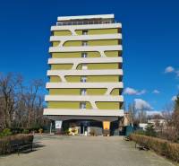 Vícepatrový bytový dům v Komárně s moderním designem, obklopený zelení a lavičkami.