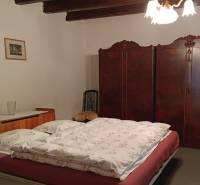 Ložnice v rodinném domě s dřevěným stropem, starožitnou skříní a klasickým lustrem.