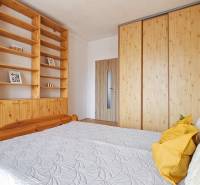 Ložnice v 2pokojovém bytě s postelí, skříní a policemi, podlaha s dřevěným dekorem.